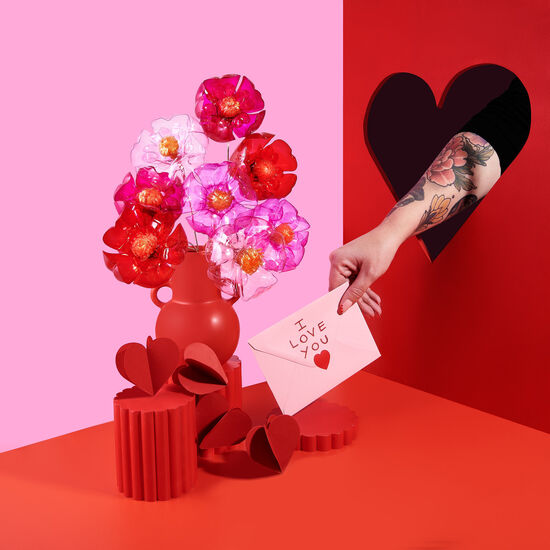 An arm with flower tattoos pokes through a heart shape cut out, holding a bouquet of pink and red recycled plastic bottle flowers