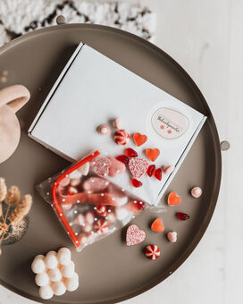 Red and white sweets pouring out of a clear cellophane bag with white spots, onto a table with our signature white gift box in the background