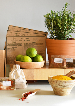 Fresh, potted thyme and recipe cards sit on a shelf, with spice packets, a bowl filled with turmeric, and limes in a dish in the foreground