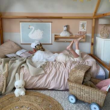 Looking in on a girl laying on her house frame bed reading. On the floor is a Miffy lamp and an Olli Ella Strolley. The wall is decorated with a wide horizontal
