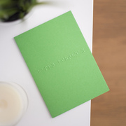 A vibrant green card lay flat on a table with happy birthday written in lower case grade one braille. There is a candle in the bottom left and a blurry plant in
