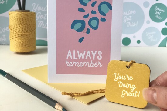 A card with a wooden bag charm attached. The charm is printed with words of encouragement You're doing great!
