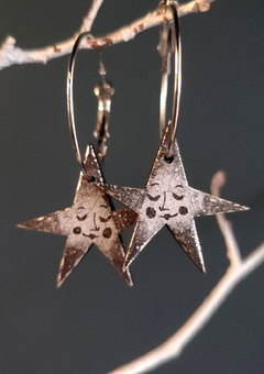 A pair of silver hoop earrings with black and grey stars with faces hanging from them. The background is dark grey and the earrings hang from white twigs. 