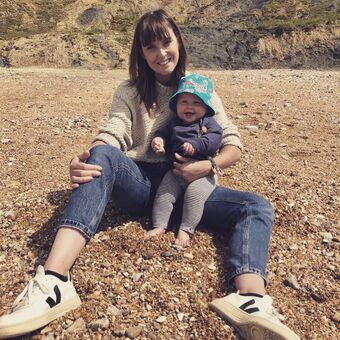  A Woman sits on a pebble beach with a baby. Both are smiling, there is a rocky cliff in the background