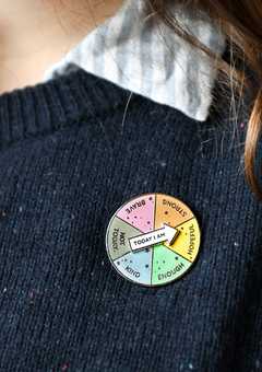A spinning enamel pin badge, with six segments each with positive words in, is pinned to someones navy blue jumper.