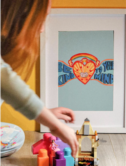 Hand painted positive quote print about mental health, featuring a heart and a rainbow on a blue background. In front a red head girl plays with lego