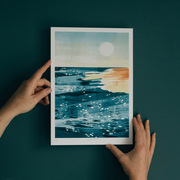 Hands holding a print of a beach scene at golden hour with a dark green wall behind. The print has gold leaf details.