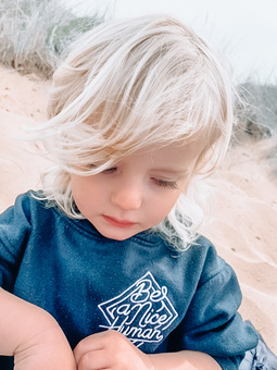 A young boy with blonde hair playing on the beach, wearing a Be a nice Human Hoodie in navy, with white embroidery