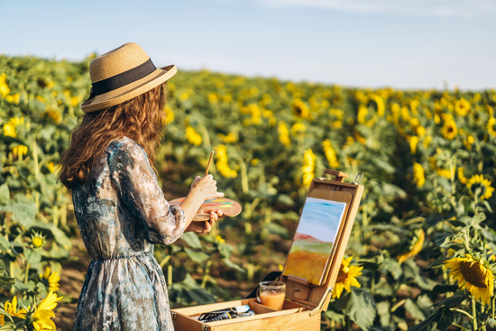 Lady in a sunflower field wearing a hat, painting on an easel with watercolours