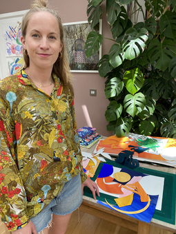 Portrait of Charlotte from Hot House Collection standing in front of her desk with art materials on. There is a big monstera plant in the background.