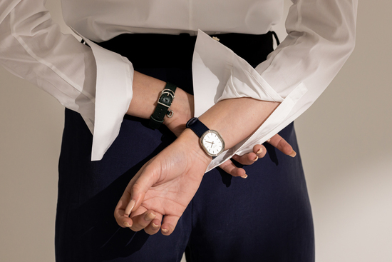 a woman seen from the back, holding her wrists behind her back. Her shirts sleeve are slightly rolled up and she has a modern watch on both wrists.