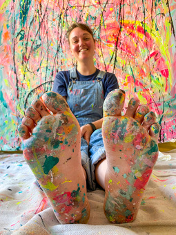 Michelle, artist and creator behind Eat Pray Pedal sitting in front of a large canvas with paint splattered feet.