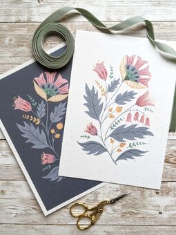 Two prints of folk art flowers in blue and pink, with a green ribbon and a pair of embroidery scissors.