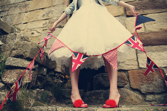 A bride wearing a denim jacket with a tea length wedding dress with red fluffy mules. The bride is holding union jack bunting as well as red gingham bunting