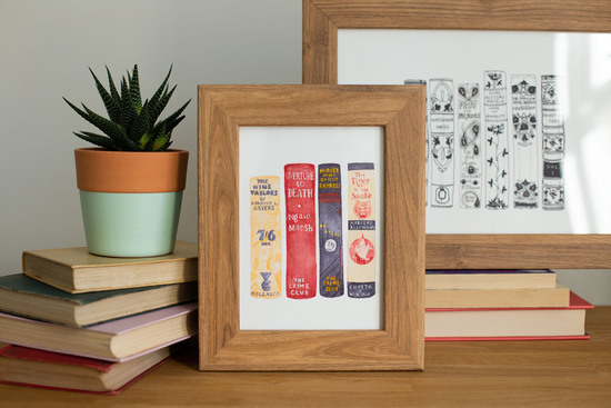 Two prints of book spines by Georgina The Librarian, in oak frames, displayed with a pile of vintage books