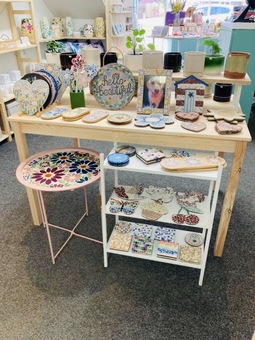 A picture of Hayleys Mosaics items in a shop. Colourful mosaic items including wreaths, photo frames and coasters are stacked on shelves 