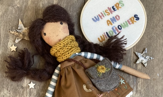 Wildflower rag doll with brown hair, mustard scarf, felt bag and illustrated brown skirt, laying on wooden background with Whiskers & Wildflowers stitched sign.