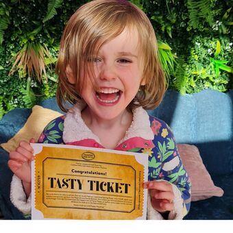 A child with blonde hair is holding a golden tasty ticket and laughing. 