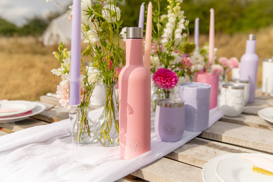 Pink Partner in Wine insulated bottle sits next to an insulated wine tumbler on a rustic table covered in flowers and candles