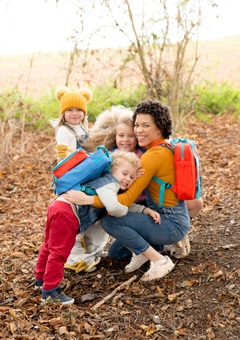 A mixed race lady cuddling three small children. They are all wearing magnetic backpacks