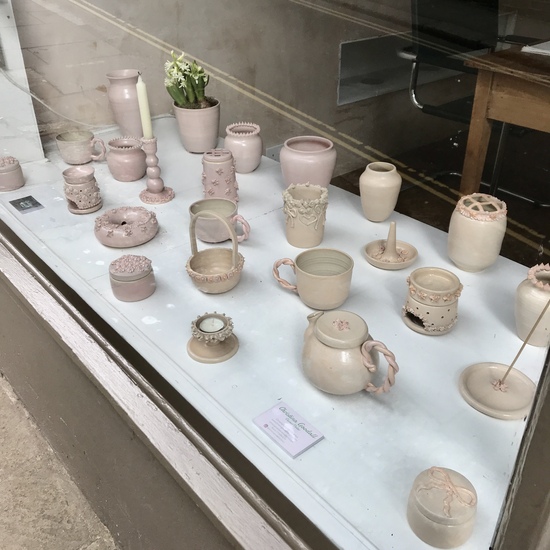 Pink and peach handmade pottery in shop window including teapot, mugs, vases, and candle holders.