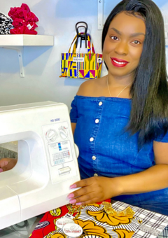 Black woman wearing a blue dress and smiling sitting down next to a sewing machine and in front of a shelf.