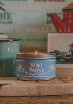 a lit wild waves candle in a tin, on a wooden side table