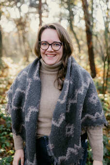 A white woman is standing in a forest, she is smiling and wearing a scarf and glasses.