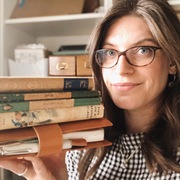 Georgina, a white woman in her 30's with dark hair and tortoiseshell rimmed glasses, holds a stack of vintage hardback books, in the background is a bookshelf