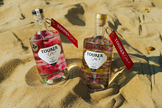 Tourer gin both flavours includes watermelon gin and mango gin bottles on the beach with sand around the base.