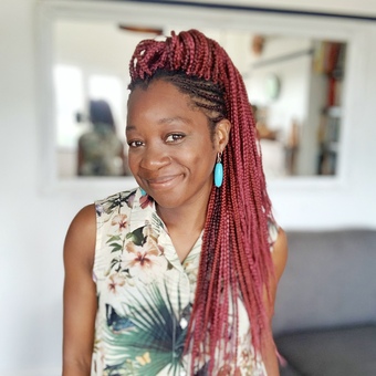A black woman with red braids, turquoise dangly earrings, wearing a sleeveless floral top. Standing in front of a mirror and smiling