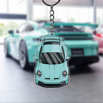 A blue car keyring with a porsche sports car on it. there is another porsche car in the background.