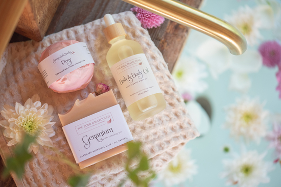 All natural The Eden Collections wellness products, bath bomb, soap and body oil are laying on a cotton towel in a beautiful bathroom with flower decoration. 