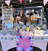 Hayley is standing behind a market stall with a pink table cloth and dark pink logo tablecloth with pretty mosaic items on display 