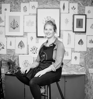 Printmaker Natalie Hallett sitting in her studio based in Devon in front of her working wall of botanical prints