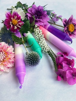 Four candles in purples, pinks and greens are positioned in a fan in amongst purple flowers.