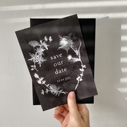 A deep inky black save the date card with white flowers, reminiscent of a photograms.