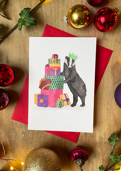 A hand drawn illustration on a greeting card of a festive badger wearing a paper Christmas hat and standing besides a pile of colourful Christmas presents