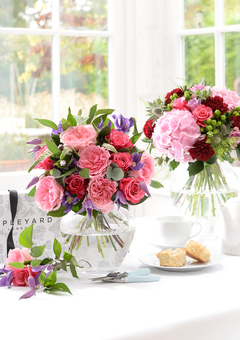 Two luxury flower bouquets in vases on a white table. The bouquets are mostly pink and include roses hydrangeas carnations and clematis