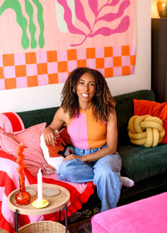 Mixed race woman with curly brown hair, sat on a green sofa with colourful cushions and graphic backdrop