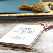 A greeting card that reads 'Thanks for being my family' lies on a table in front of other stationery items.