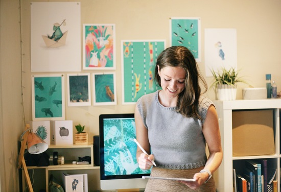 Georgia Camden stands in front of her work desk and a wall full of her art prints working on her Ipad Pro.