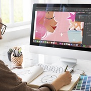 Kitty Strand working on a digital illustration at a white desk with an iMac computer