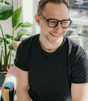 The image shows a smiling man with short brown hair and black rectangular glasses. He is wearing a black t-shirt and sitting in a brightly lit room. 