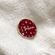 An enamel pin badge with the words I Believe in gold rests on pink fabric.