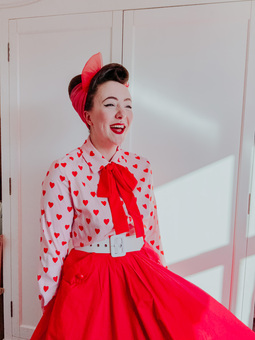 Ellie, owner of Dottie's Paper Co. is dressed is a vintage style outfit with a pink and red heart blouse, red swing skirt and hair done up in a beehive.