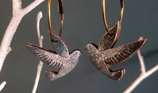 A pair of dangly earrings hanging from a white branch against a dark grey background. The earrings are blue/grey doves hanging from bronze coloured hoops