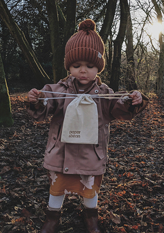 Toddler dressed in woolly hat and boots in the woodland between trees with sun rays shining through. Toddler is holding a Paper & Bean cotton bag.