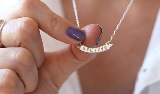 Silver banner necklace with the word 'believe' embossed. the necklace is being held and directed towards the camera.