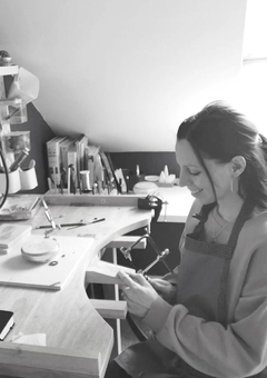 Black and white photo of jewellery designer Jodie King at a bench in a workshop. Jodie is holding a saw at the bench and is wearing an apron.